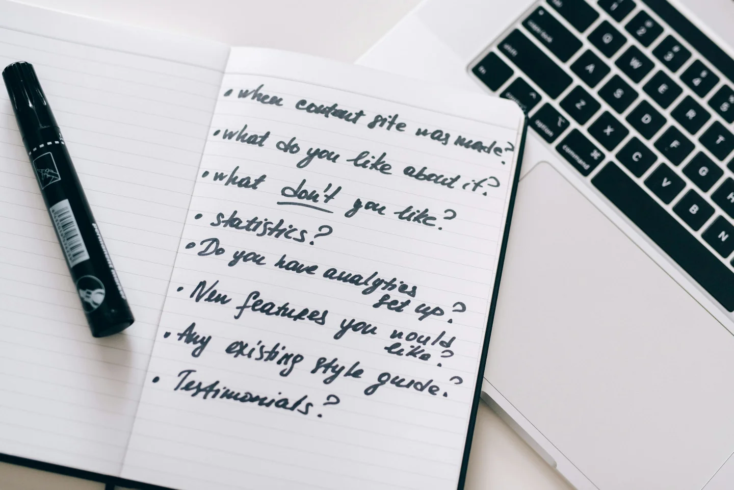 A marker on top of an open notebook with feedback questions next to a MacBook laptop